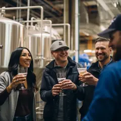Un groupe d'amis rit et trinque avec des verres de bière dans une brasserie, près d'immenses cuves en acier. Une dégustation de Camden beer.