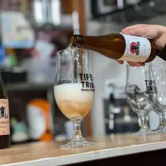 Une bière belge est versée dans un verre. Plusieurs bouteilles de bière artisanale sont alignées sur le comptoir.