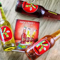 Trois bières K de Kronenbourg, une citron, une fruit rouge et une mangue, sur un fond en bois. Fraîche & fruitée ? C'est le K !