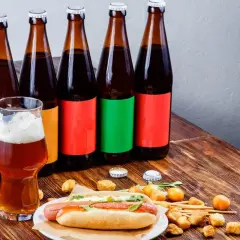 Bouteilles de bière espagnole alignées, un verre de bière ambrée et un hot-dog garni sur une table en bois.