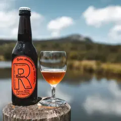 Bière trouble Rohner, artisanale et non filtrée, servie dans un verre à côté de sa bouteille, avec un lac et des montagnes en arrière-plan.