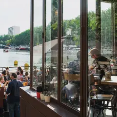 Terrasse animée au bord de l'eau, avec des gens attablés et des bateaux. Ambiance estivale à la Paname Brewing Company.