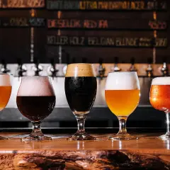 Cinq verres de bières aux couleurs variées, dont une IPA, sur un comptoir en bois. Le menu au mur liste les différentes bières.