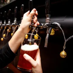 Main d'une personne servant une bière ambrée avec une mousse abondante, sous des lumières décoratives, près d'un **ballast point** de tireuses.