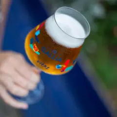 Main d'une personne tenant un verre de bière Gallia, orné de motifs colorés. La bière est dorée avec une mousse blanche.