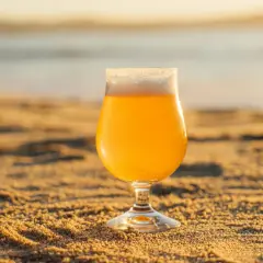 Verre de bière insolite sur le sable doré, avec la mer en arrière-plan au coucher du soleil.