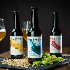 Trois bouteilles de bière artisanale Thétys : Golden Ale, Grisette et American Pale Ale. Orge et houblon frais accompagnent ces noms de bière originaux.