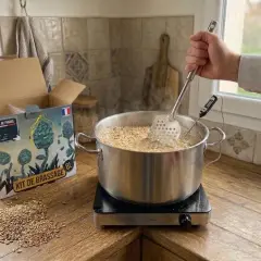 Kit de bière blanche en fabrication. Un homme remue le moût dans une marmite avec une écumoire et un thermomètre.