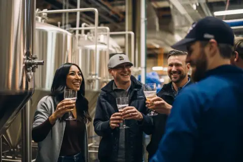 Un groupe d'amis rit et trinque avec des verres de bière dans une brasserie, près d'immenses cuves en acier. Une dégustation de Camden beer.