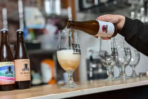 Une bière belge est versée dans un verre. Plusieurs bouteilles de bière artisanale sont alignées sur le comptoir.