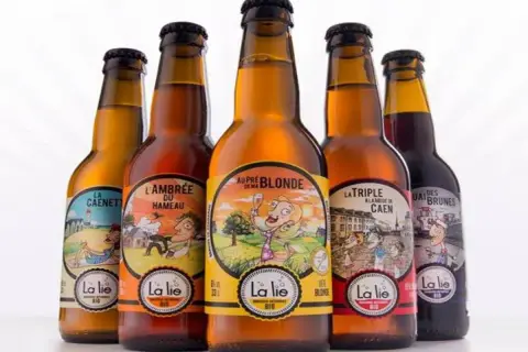 Cinq bouteilles de bière artisanale "La Lie", dont la meilleure bière normande. Des étiquettes colorées illustrent des scènes champêtres et urbaines.