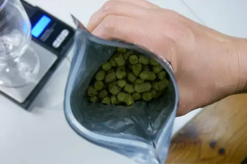 Main d'un brasseur tenant un sachet de houblon, prêt à créer une bière aromatique. Une balance et un verre sont visibles.