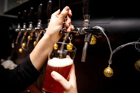 Main d'une personne servant une bière ambrée avec une mousse abondante, sous des lumières décoratives, près d'un **ballast point** de tireuses.