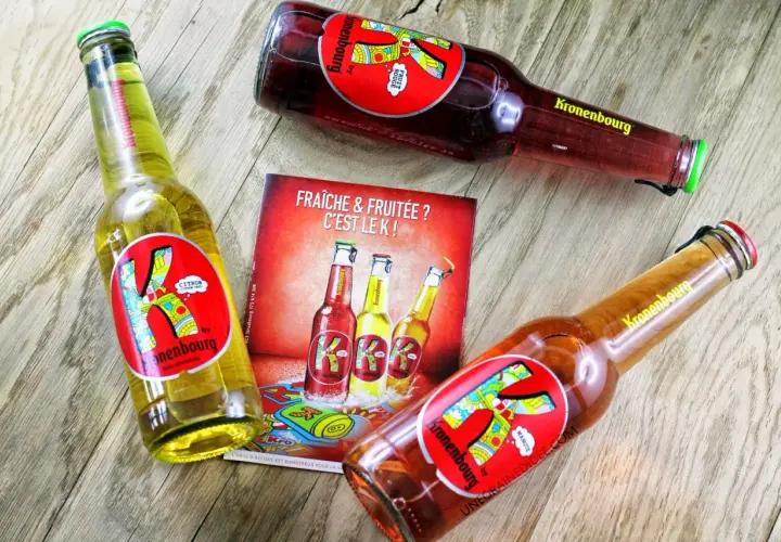 Trois bières K de Kronenbourg, une citron, une fruit rouge et une mangue, sur un fond en bois. Fraîche & fruitée ? C'est le K !