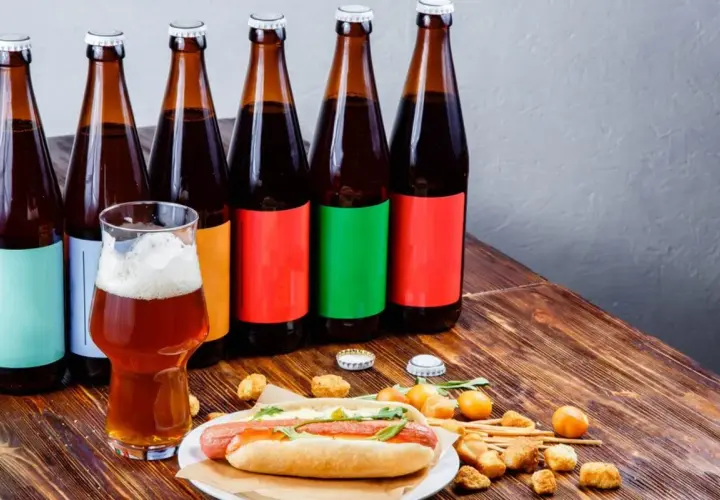 Bouteilles de bière espagnole alignées, un verre de bière ambrée et un hot-dog garni sur une table en bois.