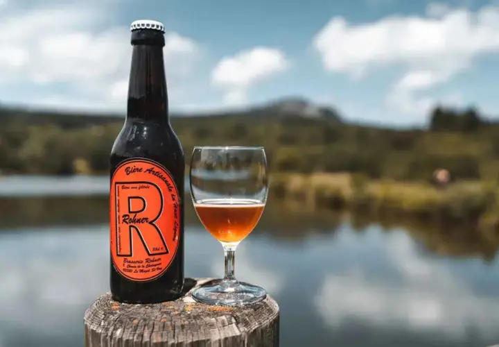 Bière trouble Rohner, artisanale et non filtrée, servie dans un verre à côté de sa bouteille, avec un lac et des montagnes en arrière-plan.