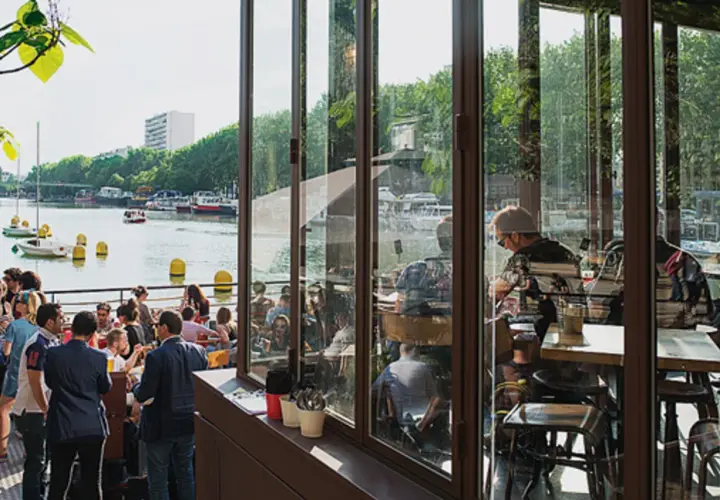 Terrasse animée au bord de l'eau, avec des gens attablés et des bateaux. Ambiance estivale à la Paname Brewing Company.