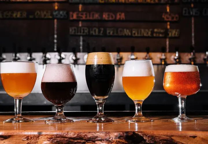 Cinq verres de bières aux couleurs variées, dont une IPA, sur un comptoir en bois. Le menu au mur liste les différentes bières.