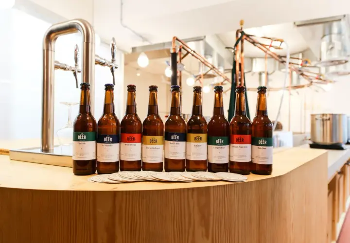 Huit bouteilles de bière artisanale alignées, chacune avec une étiquette colorée indiquant son nom et le degré alcool. Un tireuse à bière est visible à gauche.