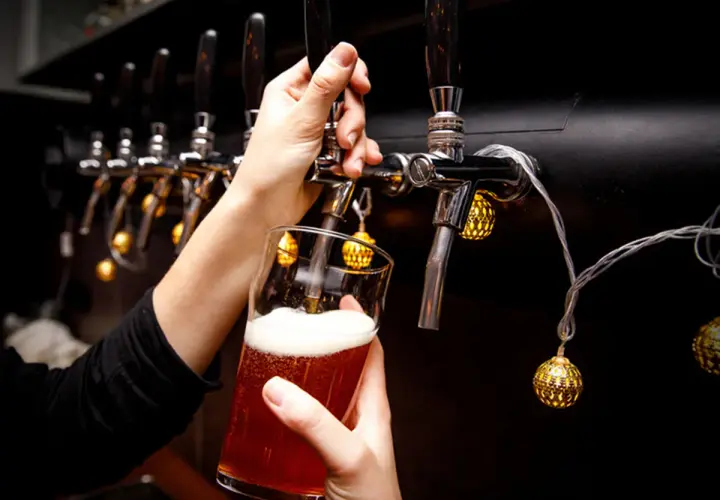 Main d'une personne servant une bière ambrée avec une mousse abondante, sous des lumières décoratives, près d'un **ballast point** de tireuses.