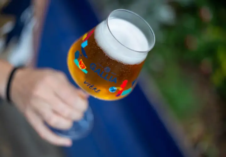 Main d'une personne tenant un verre de bière Gallia, orné de motifs colorés. La bière est dorée avec une mousse blanche.