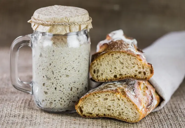 Un bocal de levain pétillant à côté de pains artisanaux coupés. Une invitation à remplacer levure par levain pour un goût authentique.