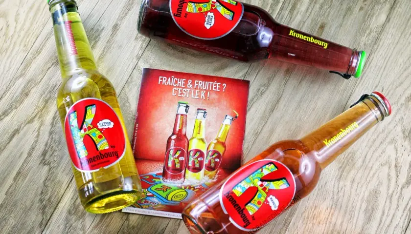 Trois bières K de Kronenbourg, une citron, une fruit rouge et une mangue, sur un fond en bois. Fraîche & fruitée ? C'est le K !