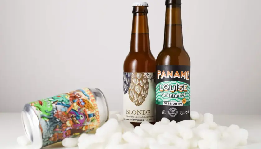 Deux bouteilles de bière et une canette colorée, dont une "Session IPA", reposent sur des flocons blancs.