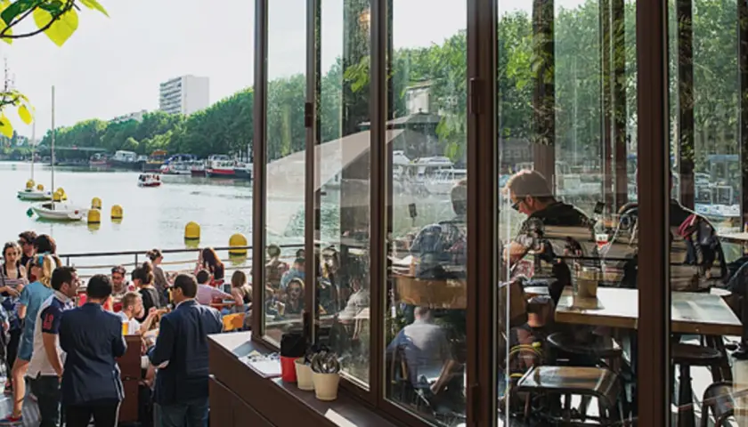 Terrasse animée au bord de l'eau, avec des gens attablés et des bateaux. Ambiance estivale à la Paname Brewing Company.