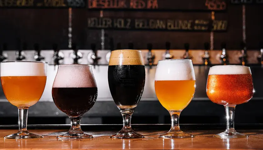 Cinq verres de bières aux couleurs variées, dont une IPA, sur un comptoir en bois. Le menu au mur liste les différentes bières.
