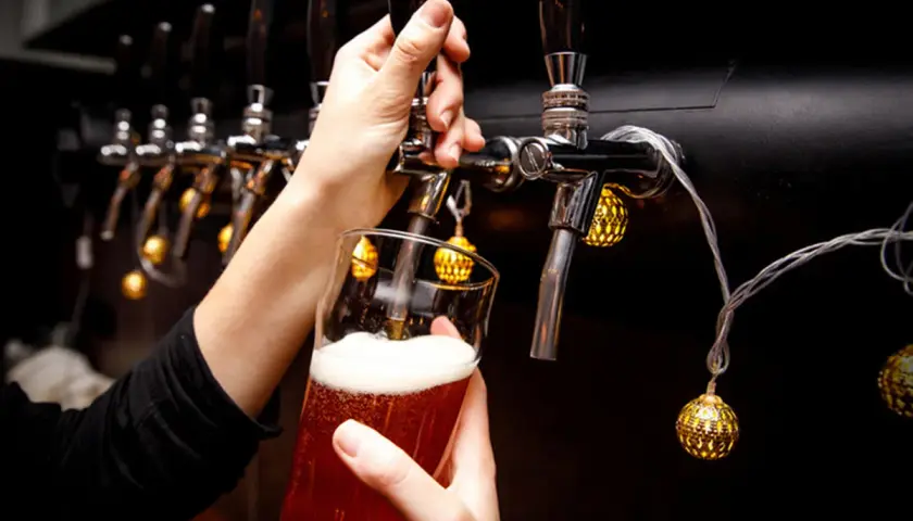 Main d'une personne servant une bière ambrée avec une mousse abondante, sous des lumières décoratives, près d'un **ballast point** de tireuses.
