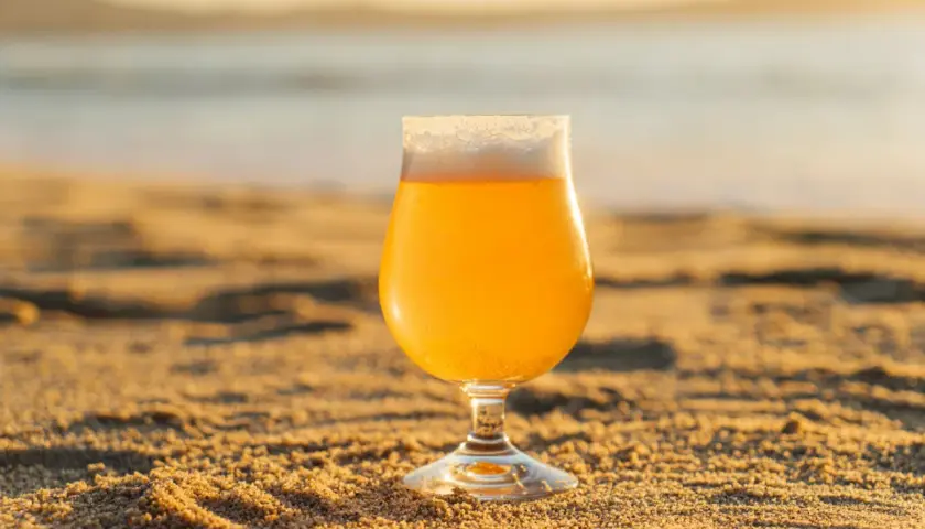 Verre de bière insolite sur le sable doré, avec la mer en arrière-plan au coucher du soleil.