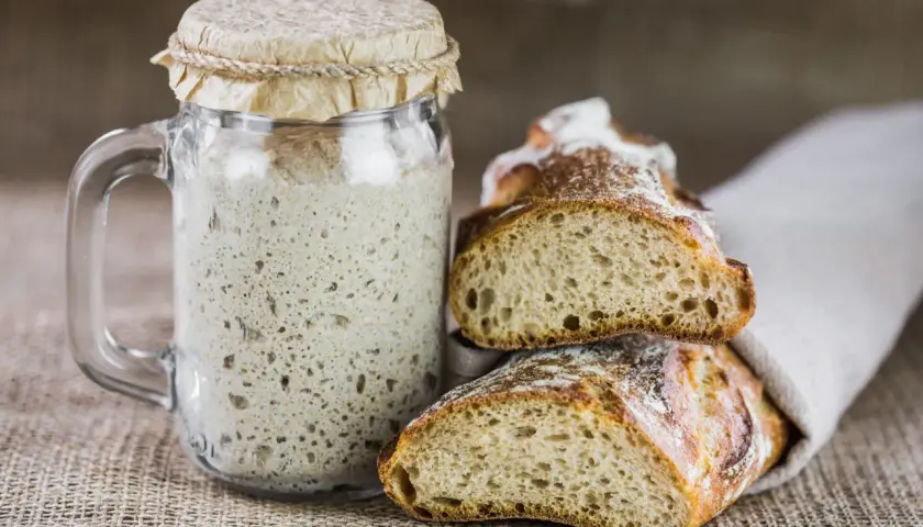 Un bocal de levain pétillant à côté de pains artisanaux coupés. Une invitation à remplacer levure par levain pour un goût authentique.