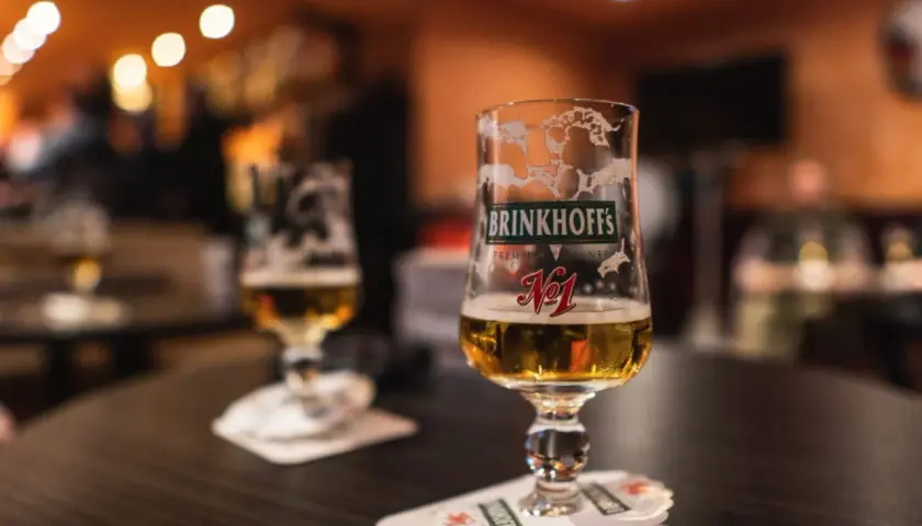 Deux verres de bières allemandes Brinkhoff's, l'un à moitié plein, sur une table sombre.