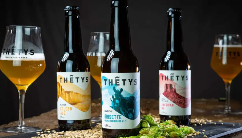 Trois bouteilles de bière artisanale Thétys : Golden Ale, Grisette et American Pale Ale. Orge et houblon frais accompagnent ces noms de bière originaux.