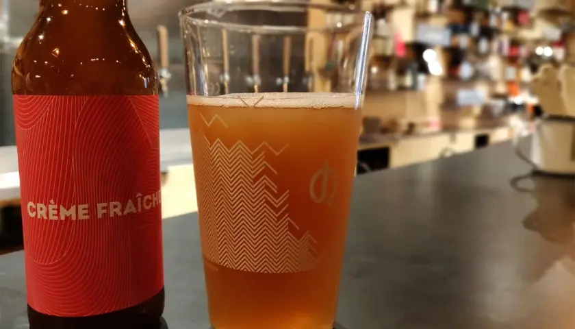 Bière artisanale "Crème Fraîche" dans un verre, avec une bouteille à côté. L'arôme subtil du houblon se marie à la douceur crémeuse.