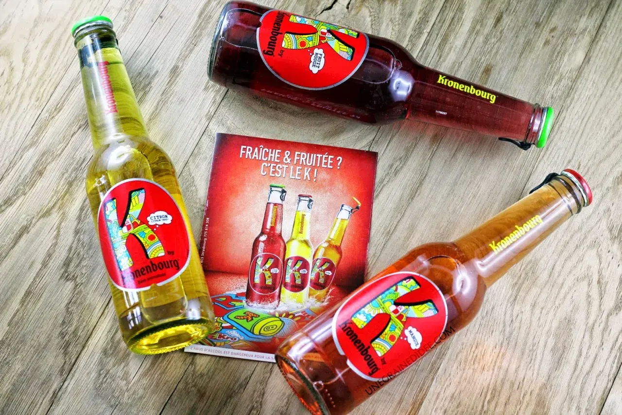 Trois bières K de Kronenbourg, une citron, une fruit rouge et une mangue, sur un fond en bois. Fraîche & fruitée ? C'est le K !