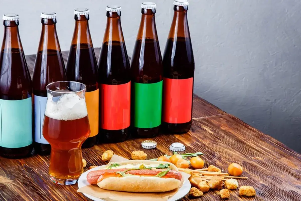 Bouteilles de bière espagnole alignées, un verre de bière ambrée et un hot-dog garni sur une table en bois.