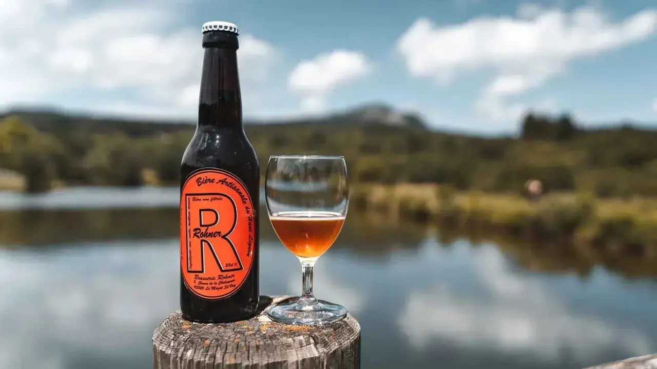 Bière trouble Rohner, artisanale et non filtrée, servie dans un verre à côté de sa bouteille, avec un lac et des montagnes en arrière-plan.