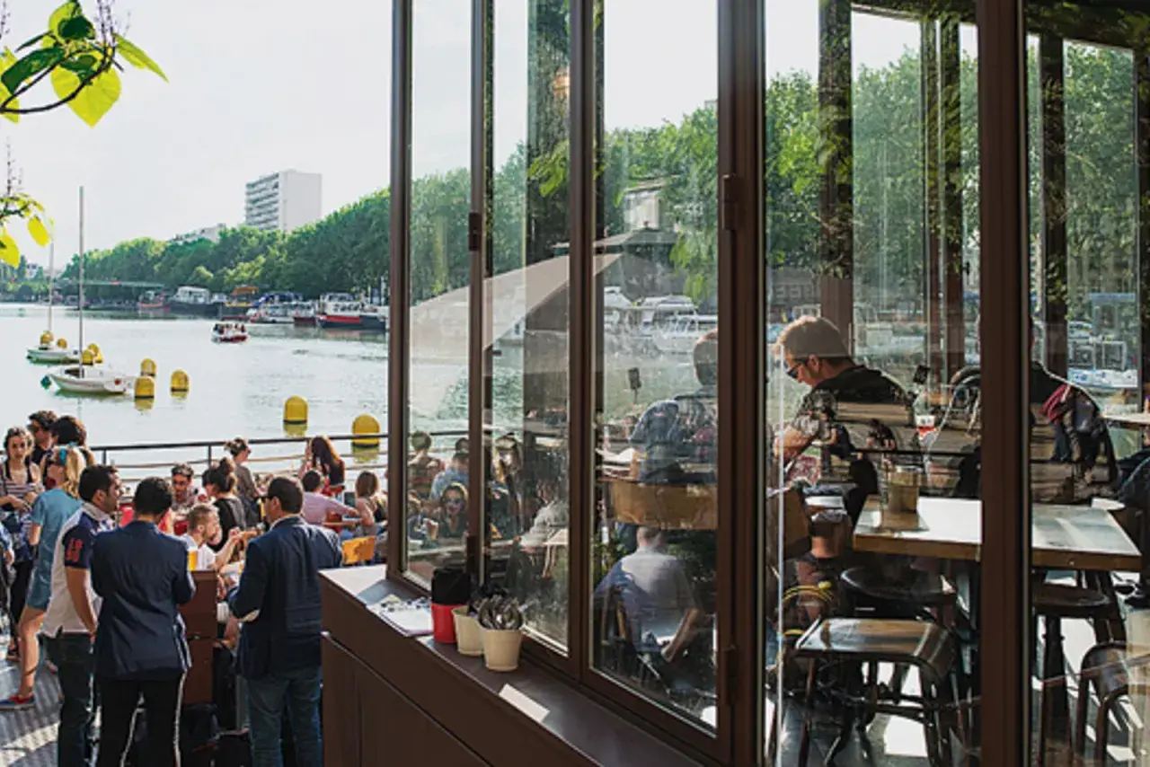 Terrasse animée au bord de l'eau, avec des gens attablés et des bateaux. Ambiance estivale à la Paname Brewing Company.