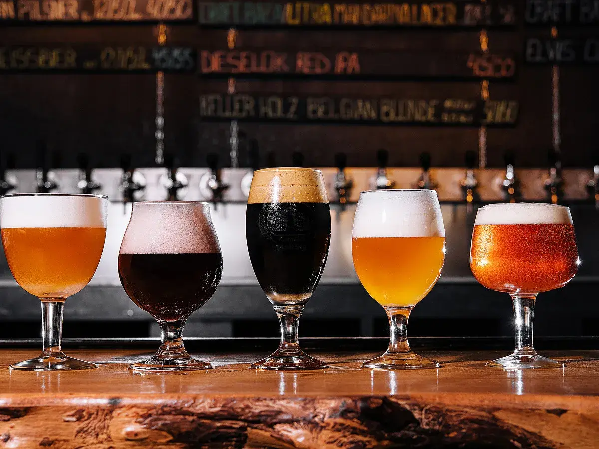Cinq verres de bières aux couleurs variées, dont une IPA, sur un comptoir en bois. Le menu au mur liste les différentes bières.