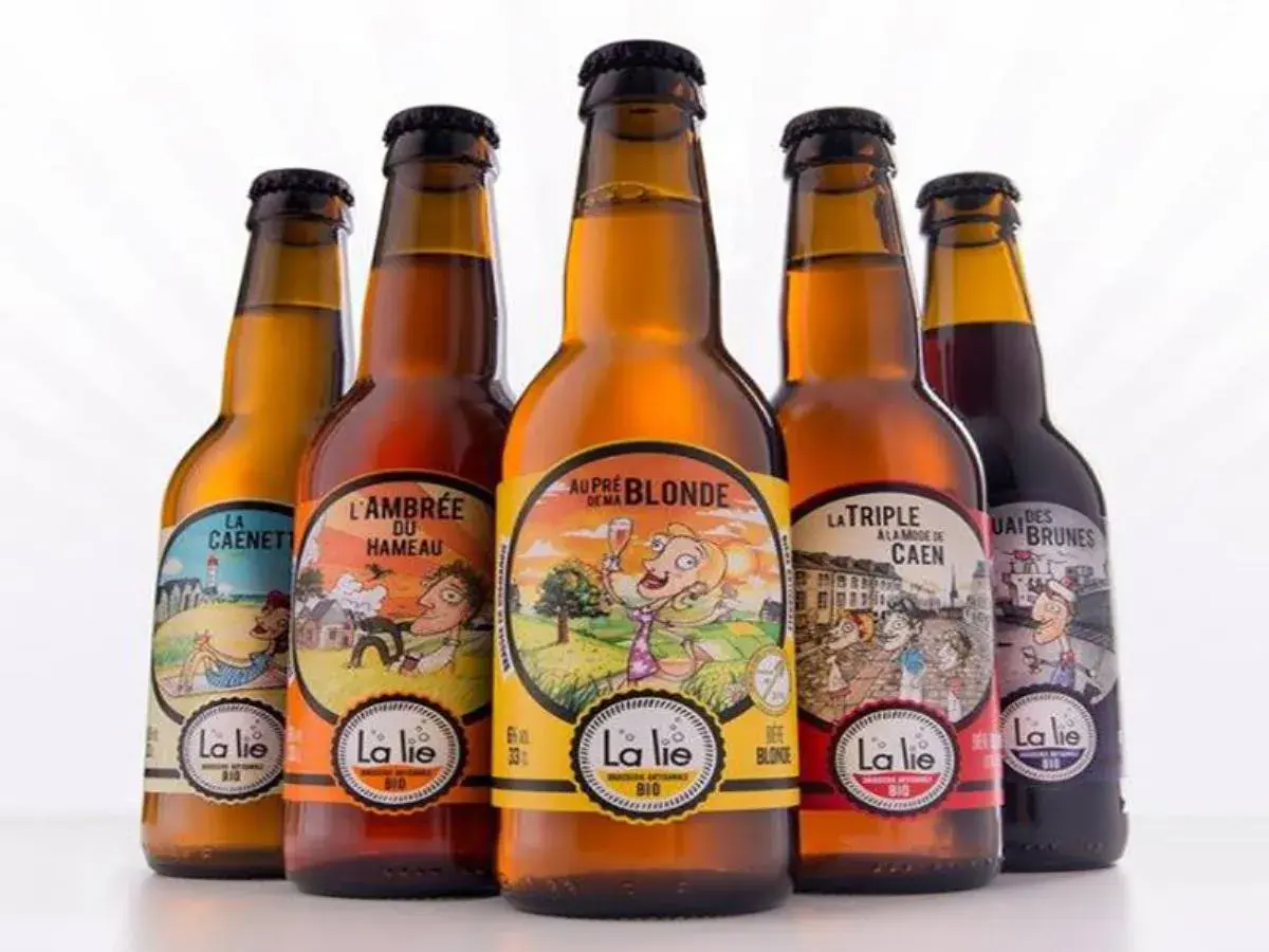 Cinq bouteilles de bière artisanale "La Lie", dont la meilleure bière normande. Des étiquettes colorées illustrent des scènes champêtres et urbaines.