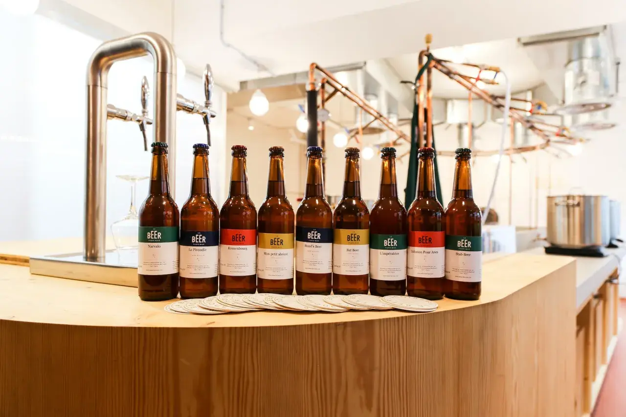 Huit bouteilles de bière artisanale alignées, chacune avec une étiquette colorée indiquant son nom et le degré alcool. Un tireuse à bière est visible à gauche.