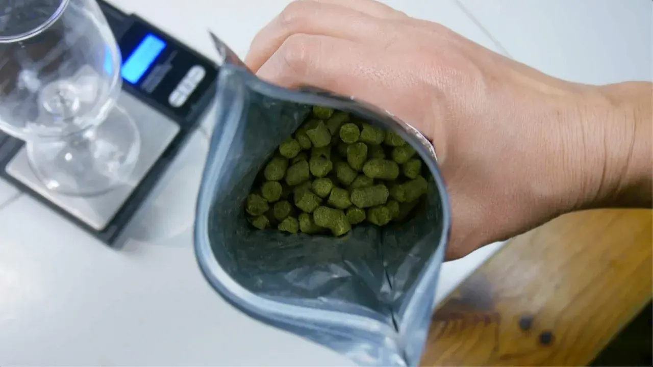 Main d'un brasseur tenant un sachet de houblon, prêt à créer une bière aromatique. Une balance et un verre sont visibles.