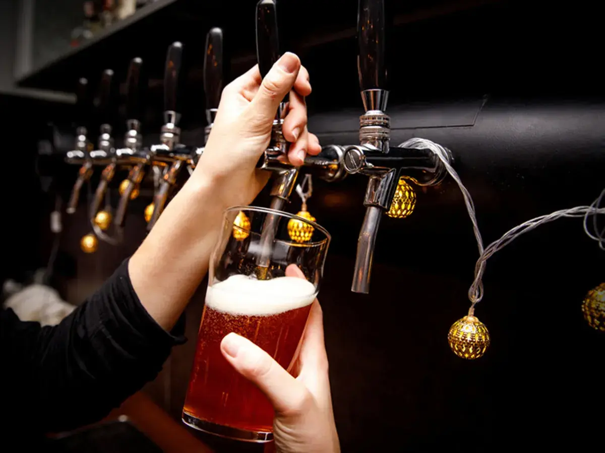 Main d'une personne servant une bière ambrée avec une mousse abondante, sous des lumières décoratives, près d'un **ballast point** de tireuses.