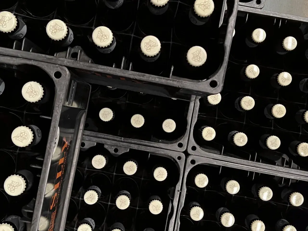 Bouteilles noires empilées dans des casiers, prêtes pour la fermentation haute. Les capsules crème arborent un motif discret.