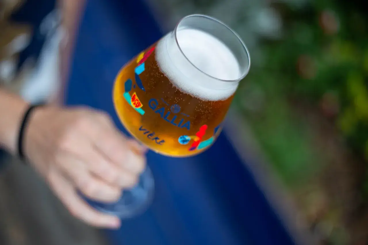 Main d'une personne tenant un verre de bière Gallia, orné de motifs colorés. La bière est dorée avec une mousse blanche.