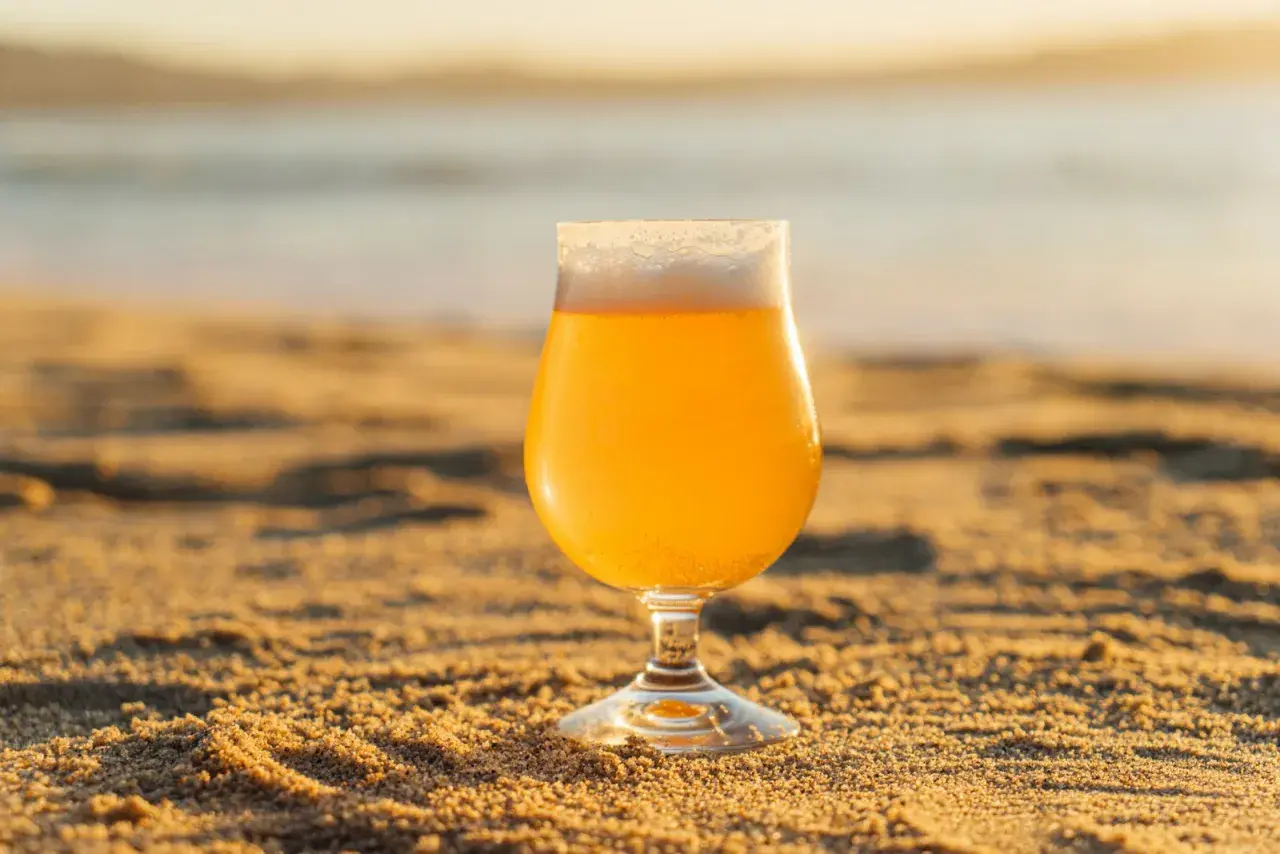 Verre de bière insolite sur le sable doré, avec la mer en arrière-plan au coucher du soleil.