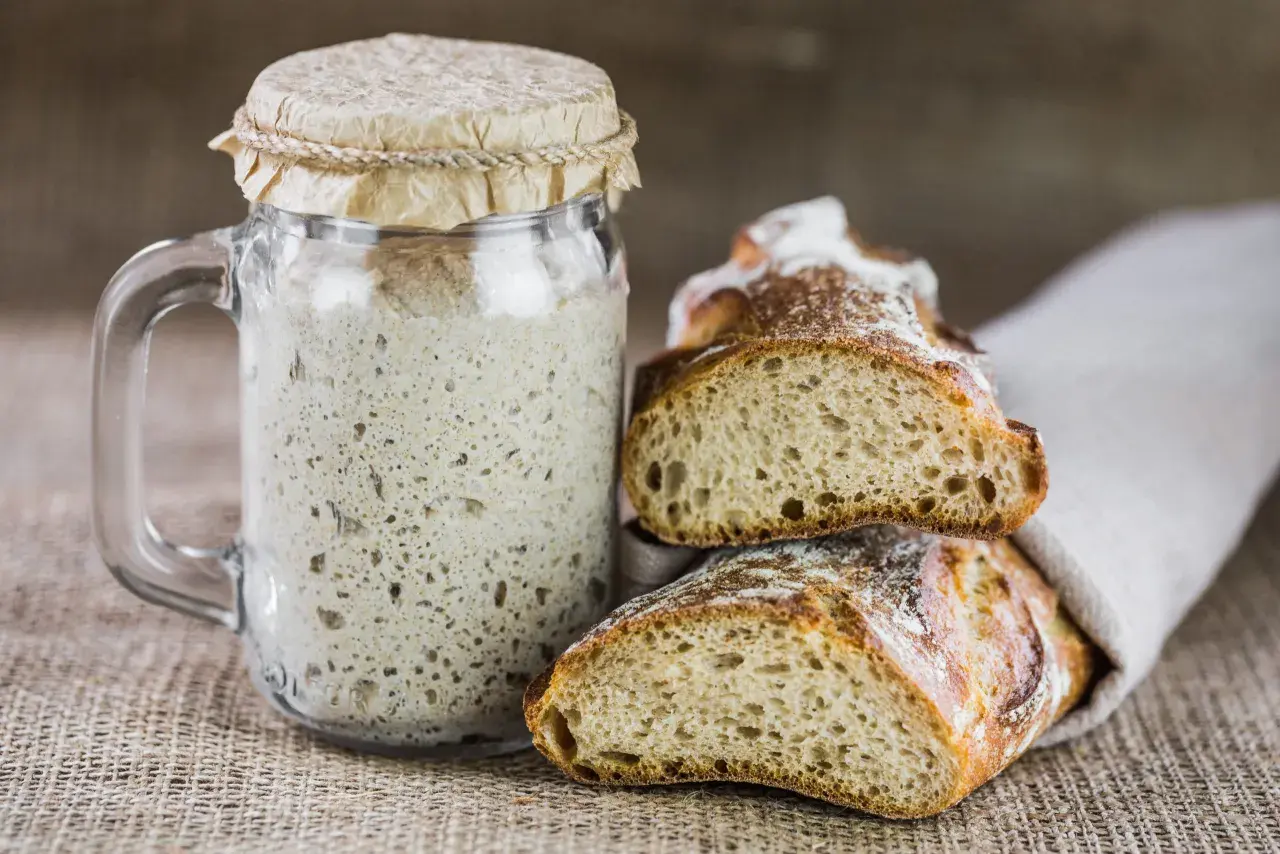 Un bocal de levain pétillant à côté de pains artisanaux coupés. Une invitation à remplacer levure par levain pour un goût authentique.