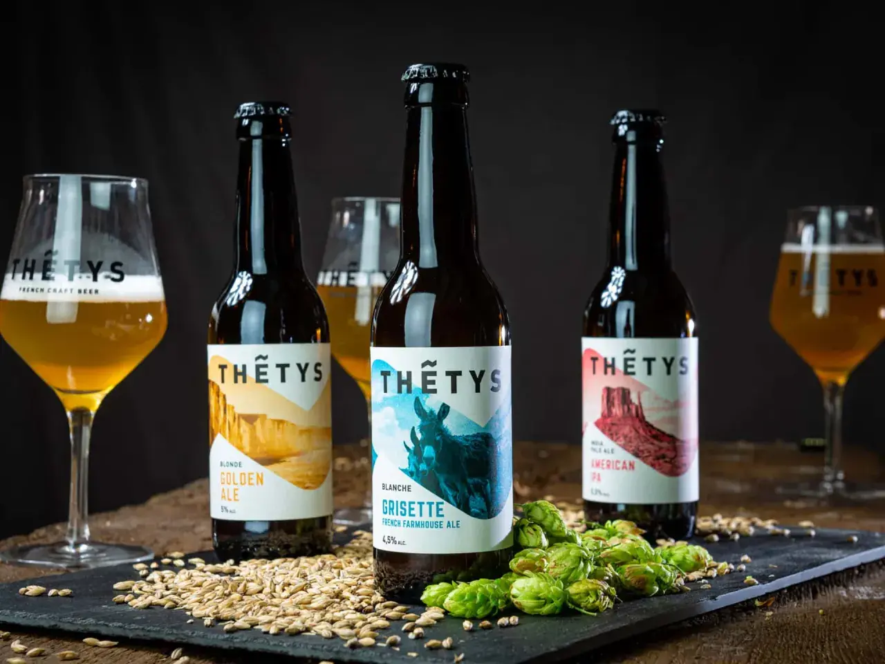 Trois bouteilles de bière artisanale Thétys : Golden Ale, Grisette et American Pale Ale. Orge et houblon frais accompagnent ces noms de bière originaux.